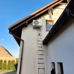 Montaż klimatyzatora na elewacji domu jednorodzinnego, widoczna drabina oparta o ścianę, jednostka zewnętrzna zamontowana pod okapem, obok okna. Na dole leży pompa próżniowa.