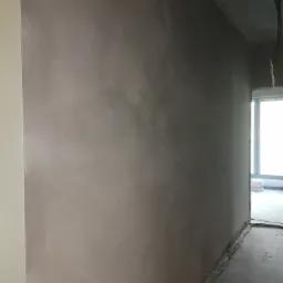 Świeżo otynkowana ściana wewnętrzna w surowym stanie, widoczne niedoskonałości i resztki materiałów budowlanych na podłodze, z fragmentem okna w tle.