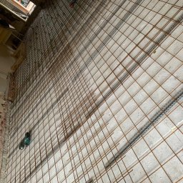 PROFESTON - Siatka zbrojeniowa ułożona na podłożu z płyt styropianowych z listwami dystansowymi, widoczna wiertarka akumulatorowa przy ścianie z odsłoniętą cegłą.