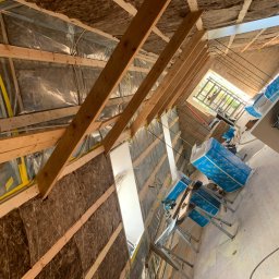 PROFESTON - Montaż izolacji termicznej z wełny mineralnej na poddaszu, widoczne drewniane krokwie i folia paroizolacyjna, częściowo oświetlone światłem dziennym wpadającym przez okno dachowe.