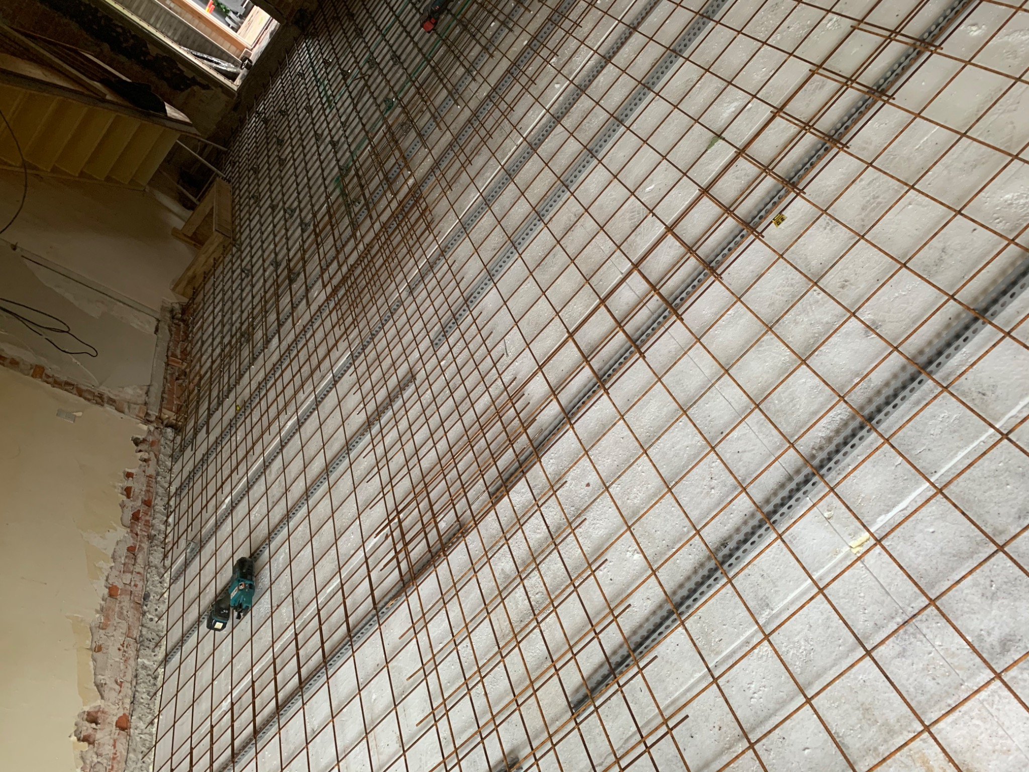 Siatka zbrojeniowa ułożona na podłożu z płyt styropianowych z listwami dystansowymi, widoczna wiertarka akumulatorowa przy ścianie z odsłoniętą cegłą.