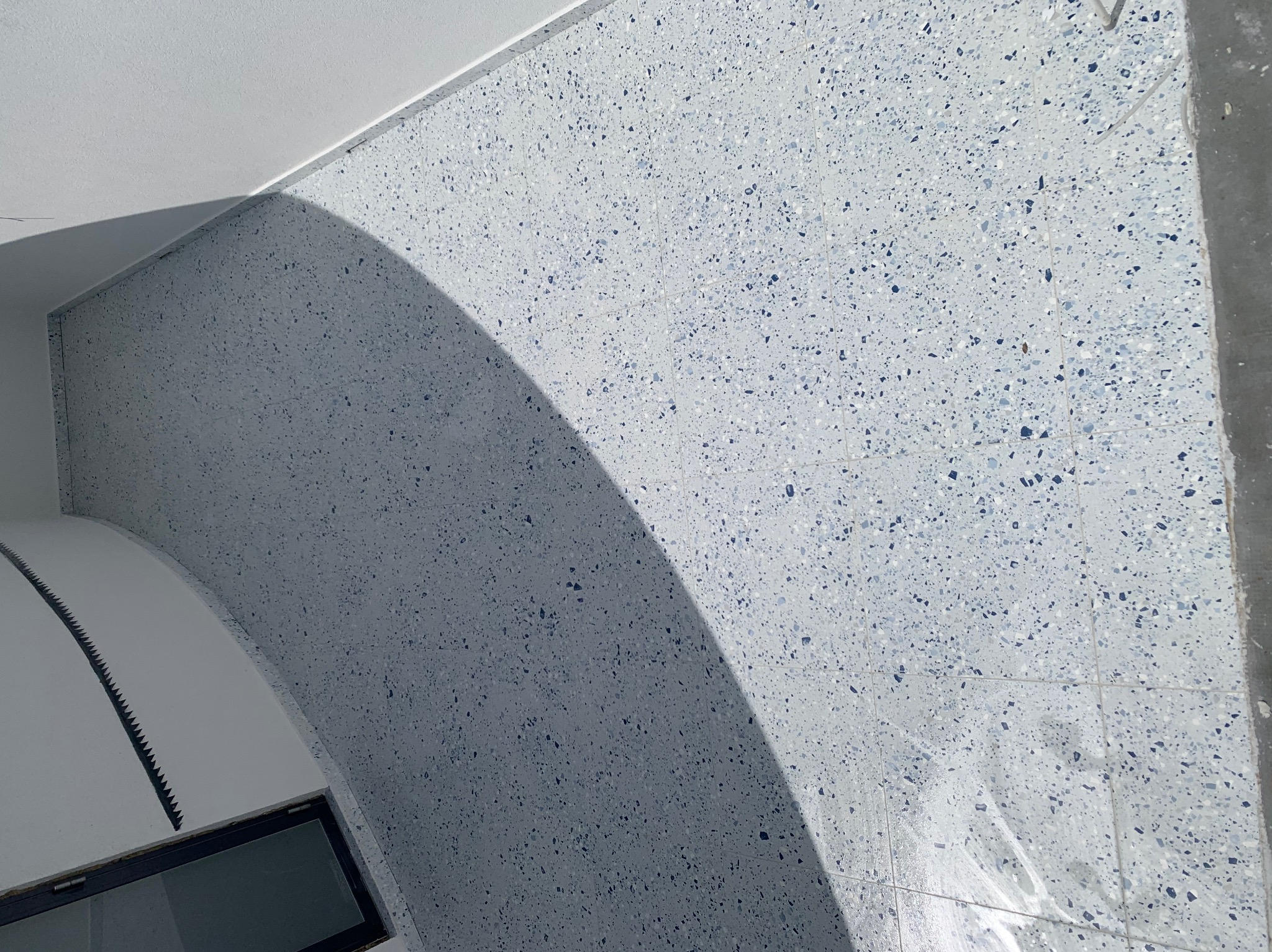 Ściana i fragment sufitu wykończone jasnoszarymi płytkami z niebieskimi i szarymi drobnymi elementami, widoczne w narożniku fragmenty okna i listwy wykończeniowej.