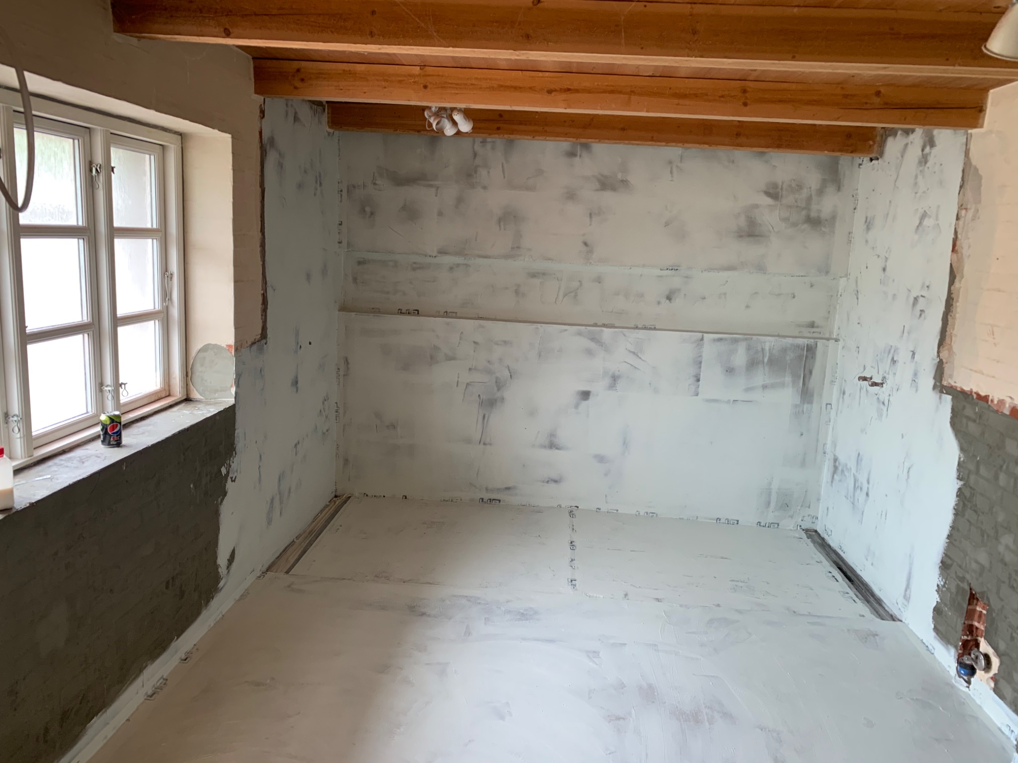 Wnętrze pokoju w trakcie remontu: ściany i podłoga pokryte świeżo nałożoną gładzią, widoczne drewniane belki stropowe, okno z napojem Pepsi na parapecie.