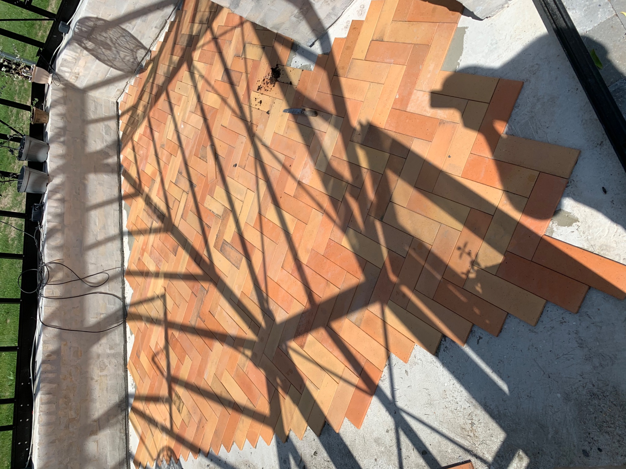 Układanie ceglanej podłogi w jodełkę w Częstochowie, widoczne ostre cienie rzucane przez konstrukcję, narzędzie leżące na cegłach i fragmenty niezakończonych prac.