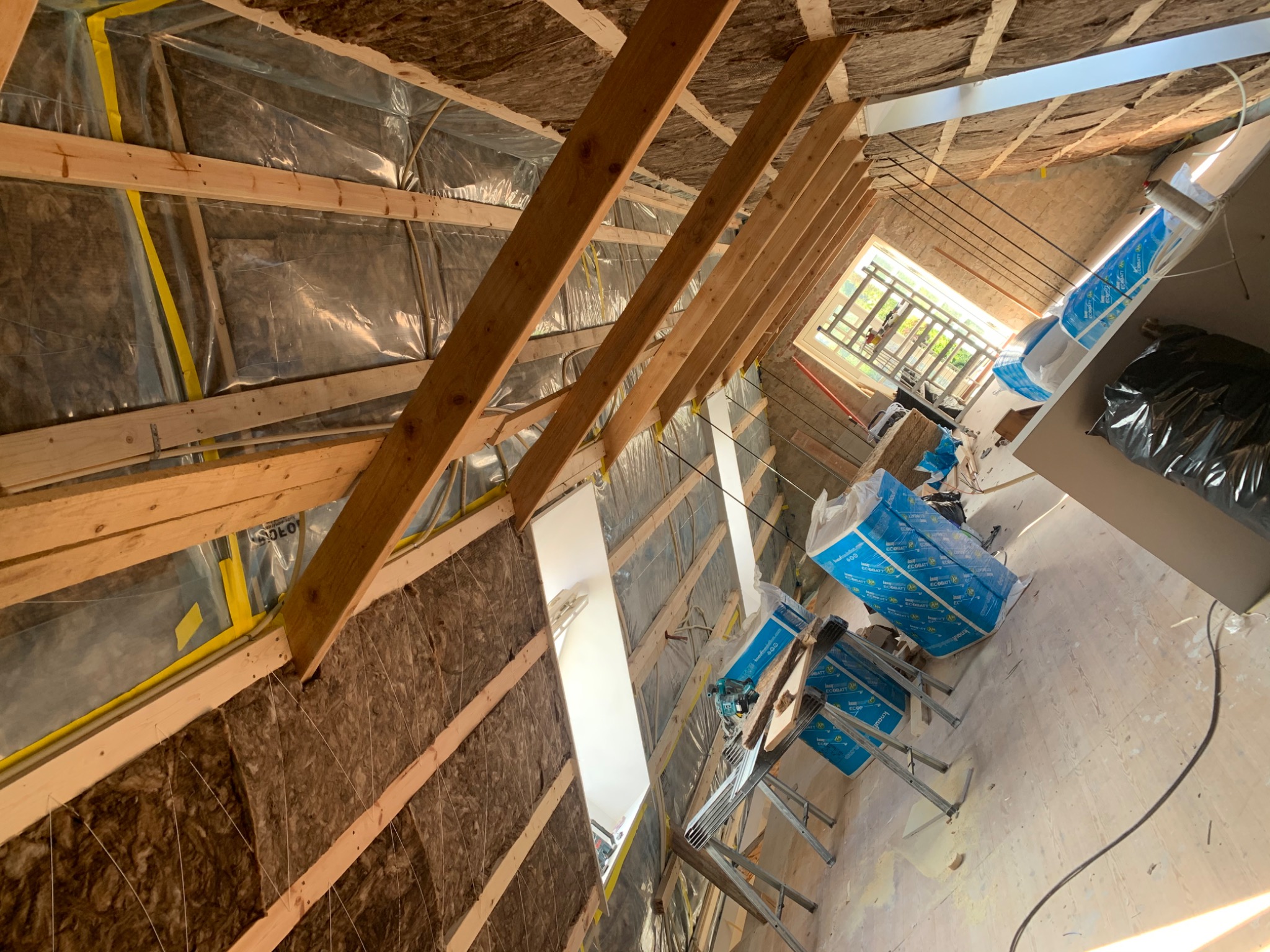Montaż izolacji termicznej z wełny mineralnej na poddaszu, widoczne drewniane krokwie i folia paroizolacyjna, częściowo oświetlone światłem dziennym wpadającym przez okno dachowe.