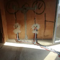 Surowe wnętrze w trakcie prac hydraulicznych: widoczne rury instalacyjne w ścianie z pustaków, zabetonowane podejścia wodne, instalacja podtynkowana, ślady po wyznaczeniu punktów na ścianie, okno...