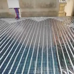 Instalacja ogrzewania podłogowego w trakcie montażu, widoczne rury grzewcze ułożone na folii izolacyjnej z siatką, surowy stan wykończenia wnętrza.