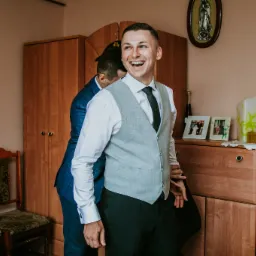 Uśmiechnięty pan młody w szarej kamizelce i czarnym krawacie, pomagający mu założyć marynarkę, w tle szafa i p&oacute;łka z pamiątkami w pokoju.