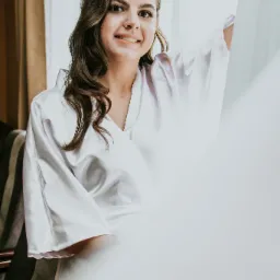 Uśmiechnięta kobieta w białym szlafroku pozuje przy oknie z delikatną firanką, włosy delikatnie kręcone, naturalny makijaż.