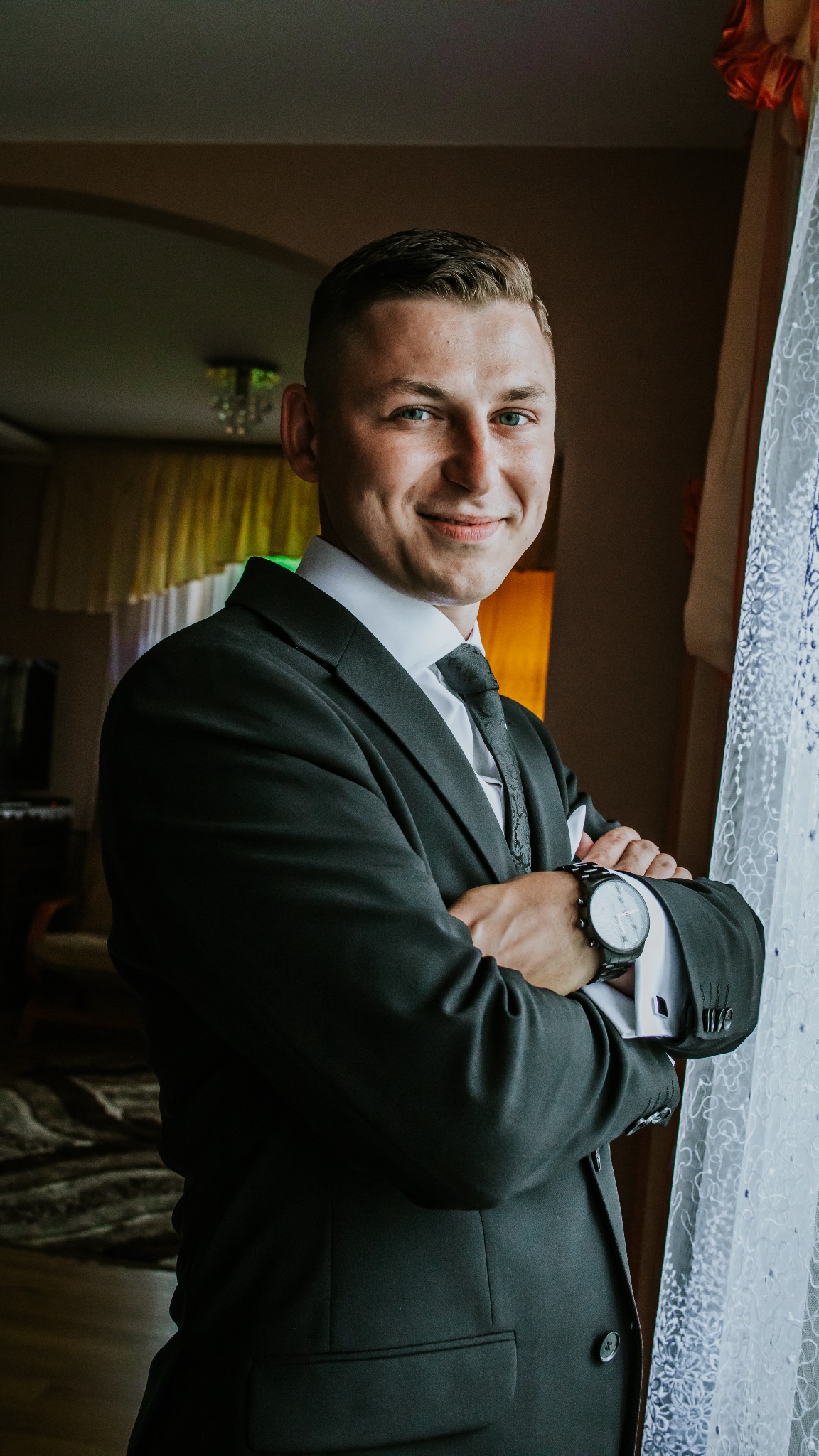 Uśmiechnięty mężczyzna w czarnym garniturze, białej koszuli i wzorzystym krawacie, stoi z założonymi rękami na tle okna z koronkową firanką, eksponując duży zegarek na nadgarstku.