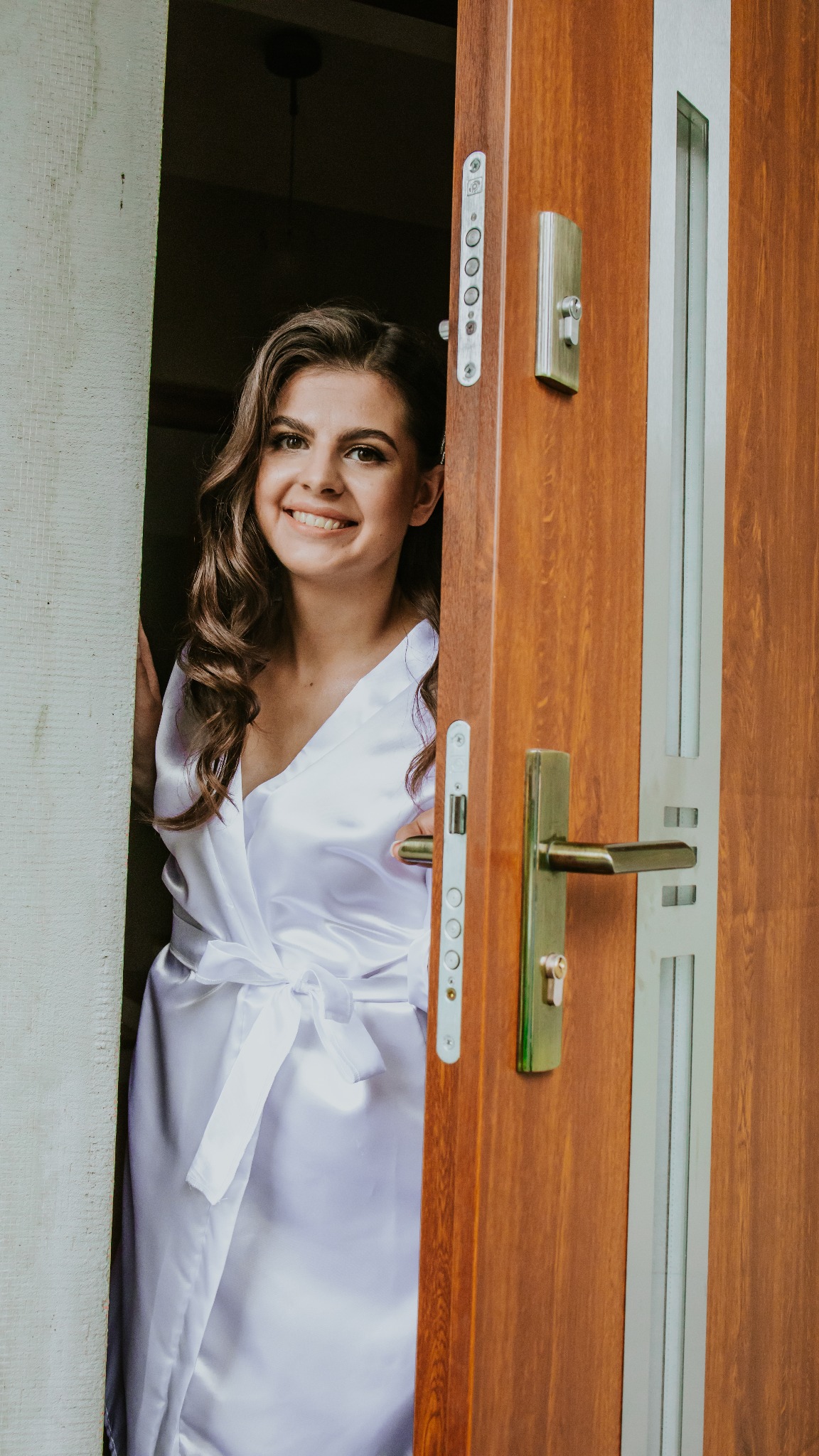 Uśmiechnięta kobieta w białym satynowym szlafroku, stojąca w drzwiach z brązowego drewna z metalowymi elementami, przygotowująca się do ślubu.
