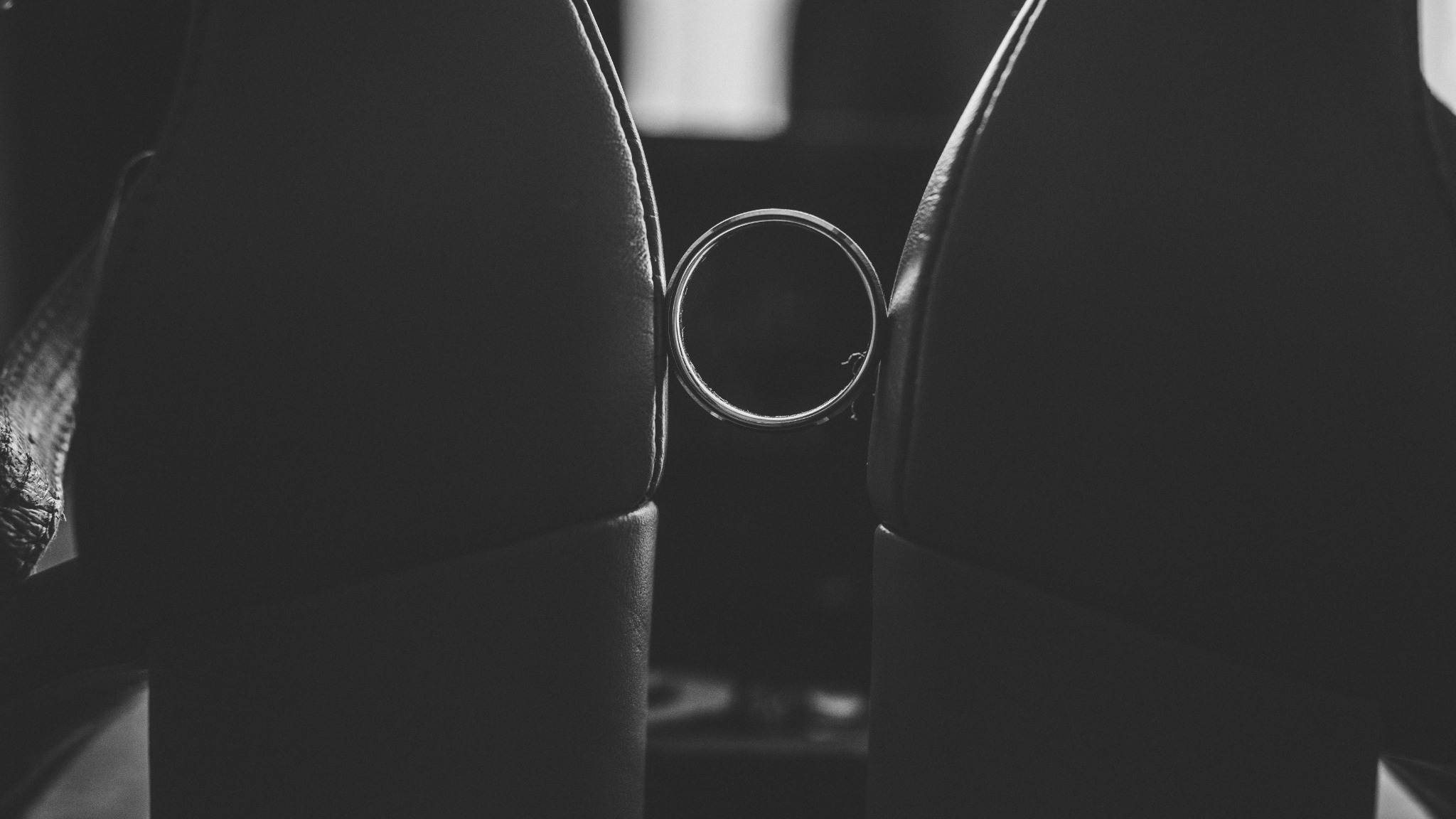 Czarno-białe zbliżenie obrączki ślubnej zawieszonej między dwoma skórzanymi oparciami krzeseł, z widoczną fakturą skóry i delikatnym światłem padającym na obrączkę.