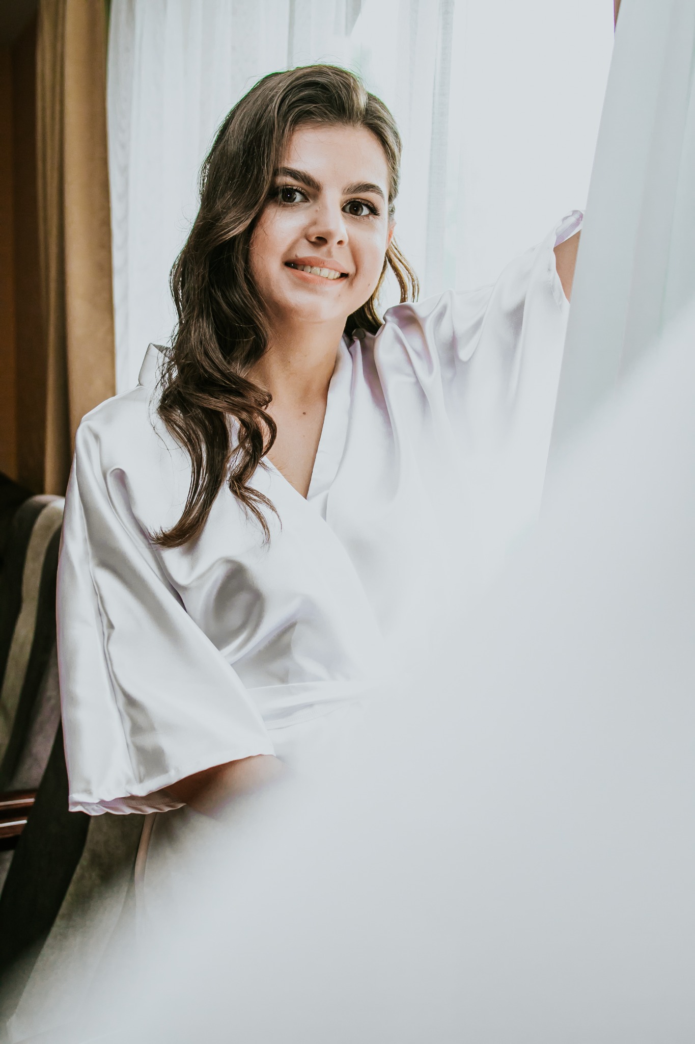 Uśmiechnięta kobieta w białym szlafroku pozuje przy oknie z delikatną firanką, włosy delikatnie kręcone, naturalny makijaż.