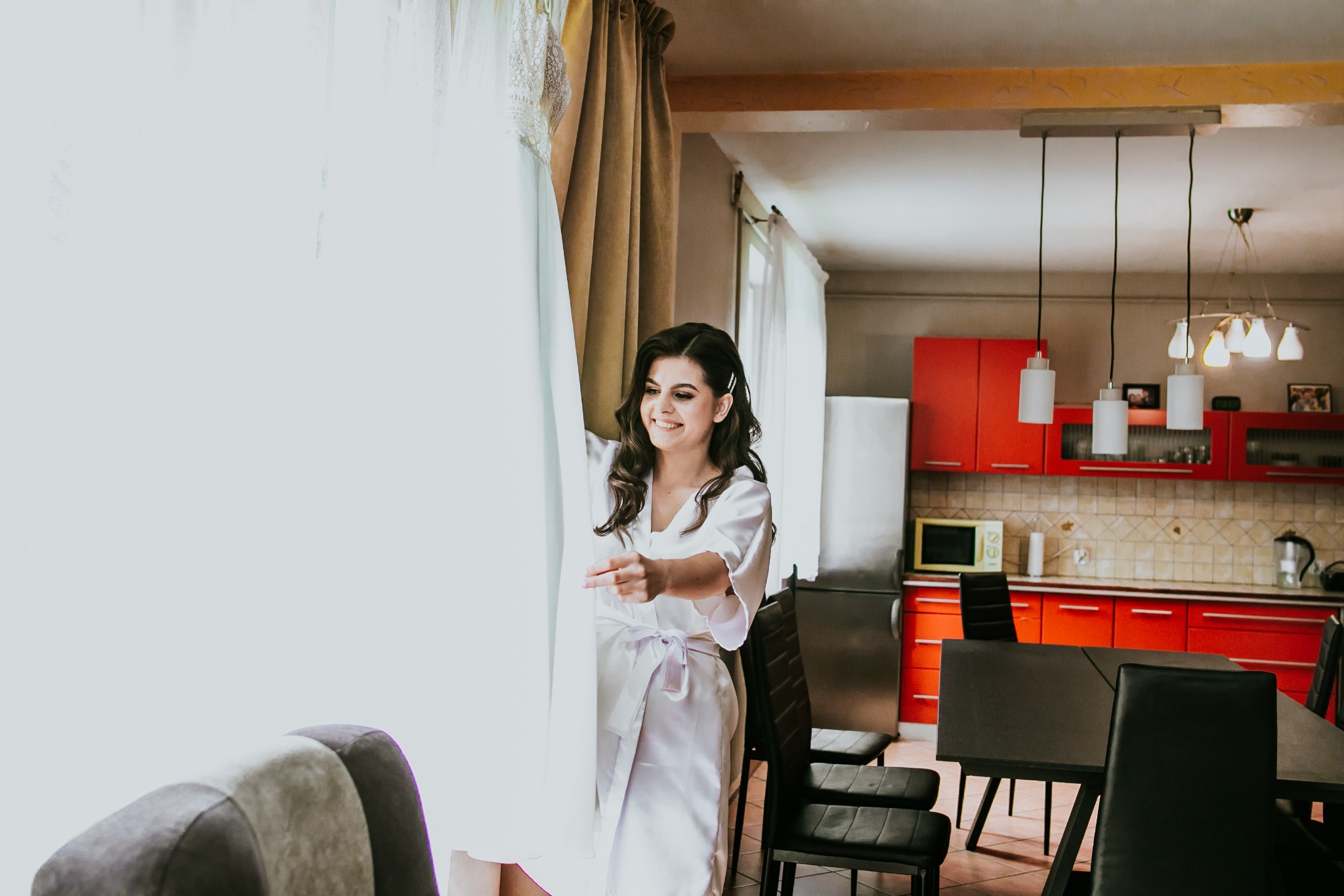 Uśmiechnięta kobieta w satynowym szlafroku, ogląda wiszącą, białą suknię ślubną z koronkowymi detalami, w tle wnętrze kuchni z czerwonymi szafkami.