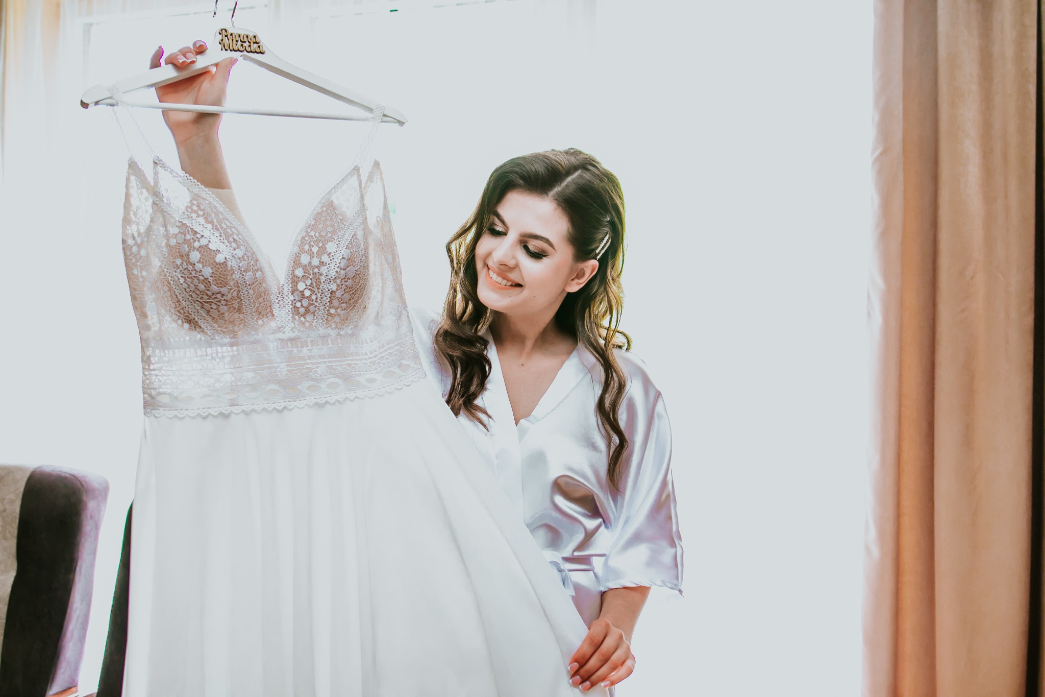 Uśmiechnięta panna młoda w satynowym szlafroku trzyma na wieszaku koronkową suknię ślubną z napisem 'Panna Młoda'.