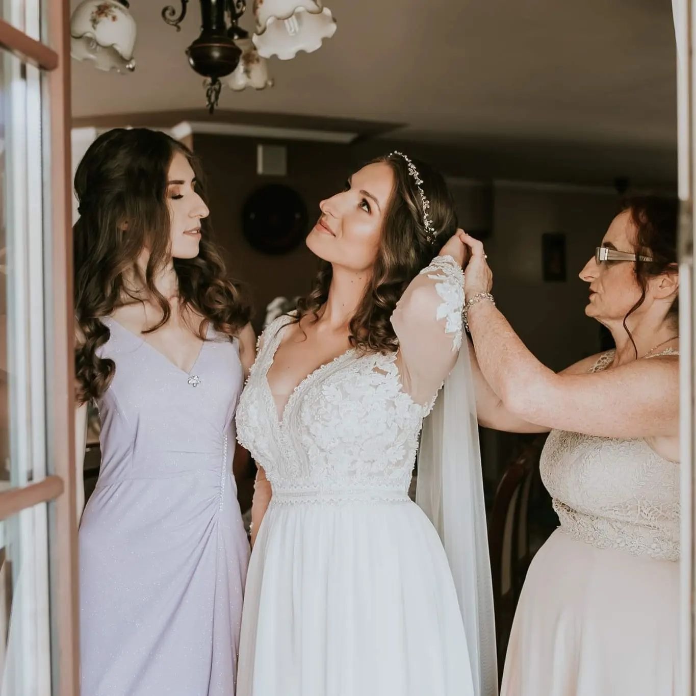 Panna młoda w koronkowej sukni spogląda w górę, podczas gdy kobieta w okularach poprawia jej welon, obok stoi druhna w liliowej sukience.