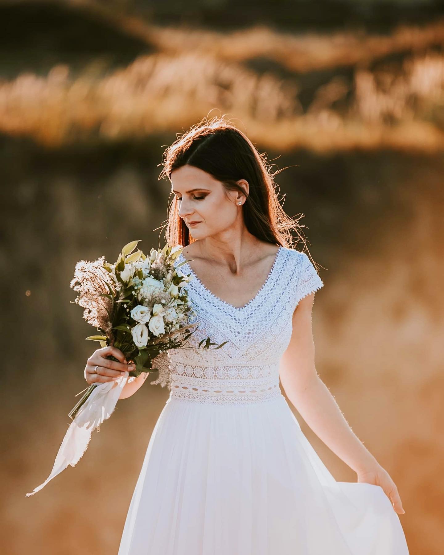 Panna młoda w koronkowej sukni, trzyma bukiet z białych róż i traw, delikatne światło zachodzącego słońca.
