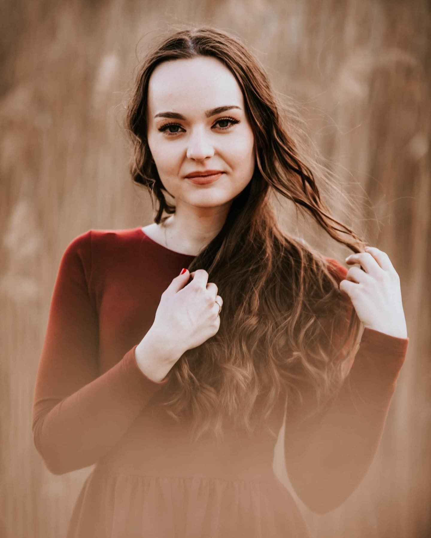 Portret kobiety w czerwonej sukience na tle suchej trawy, delikatny uśmiech, dłońmi bawi się włosami.