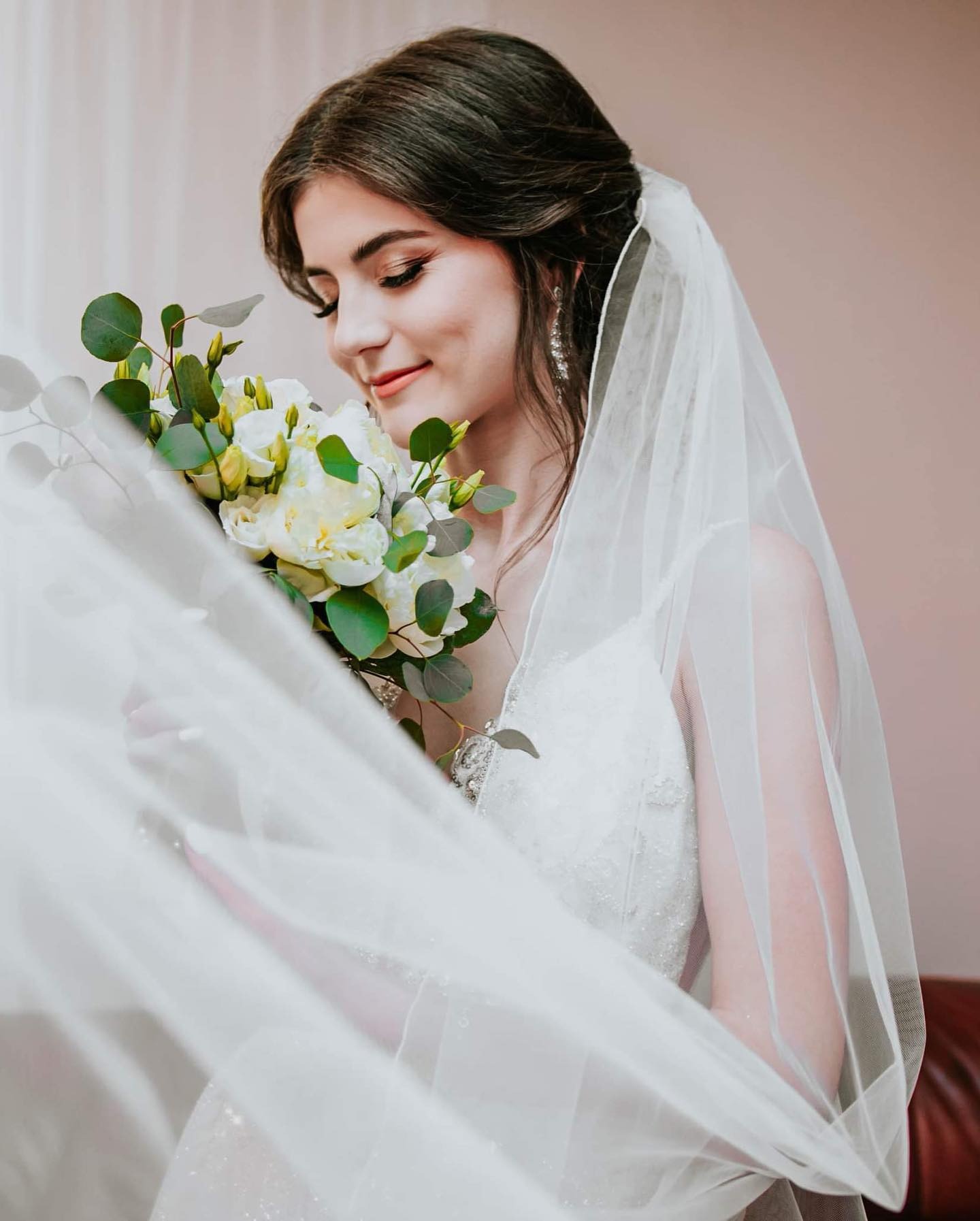 Uśmiechnięta panna młoda w białej sukni i welonie, trzyma bukiet białych kwiatów z zielonymi dodatkami, spoglądając na niego z czułością.