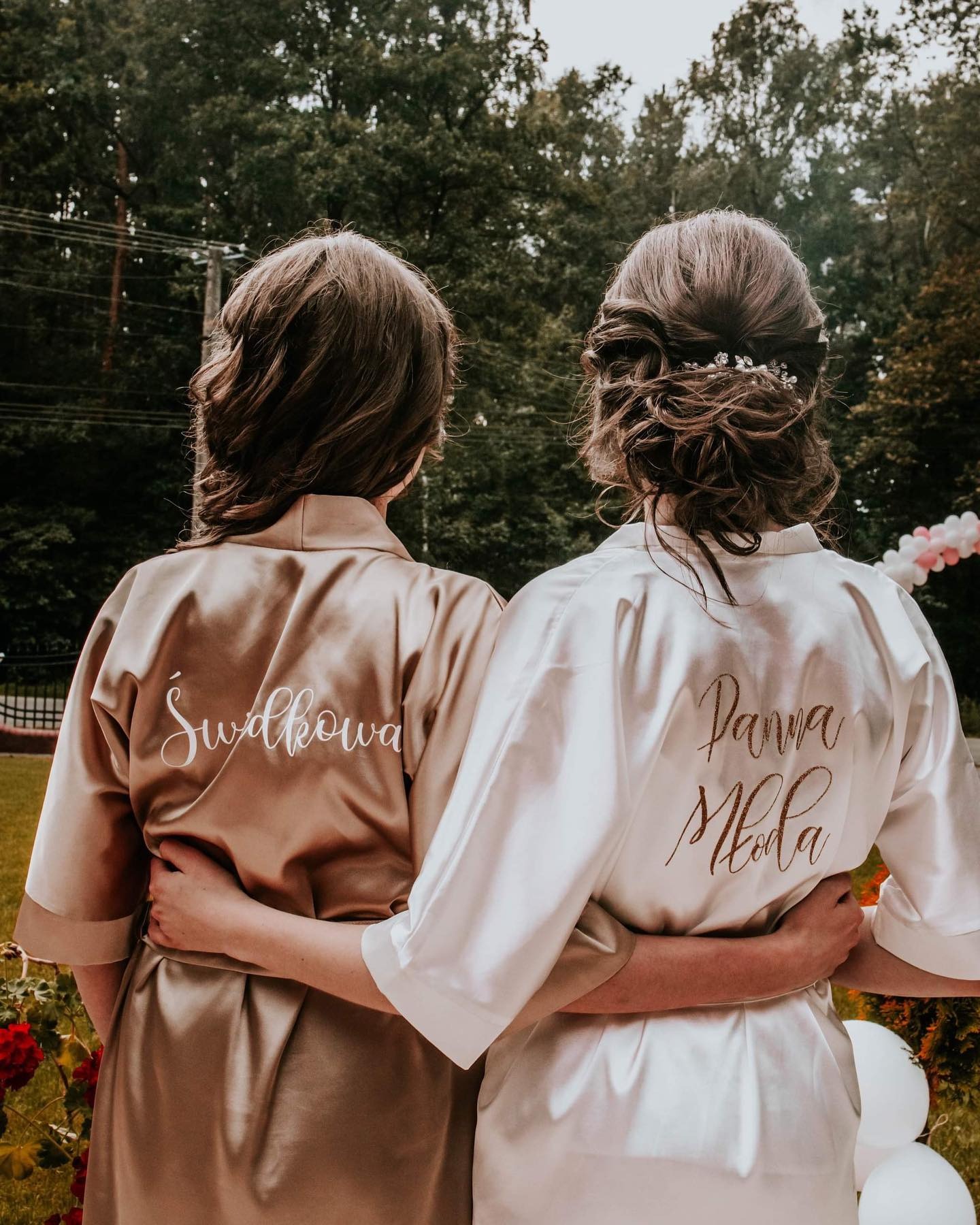 Panna Młoda i Świadkowa w satynowych szlafrokach z napisami na plecach, obejmujące się na tle zieleni i dekoracji balonowych.