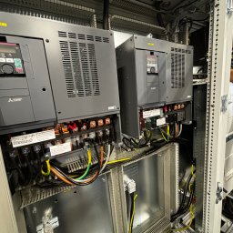 Maciej Burzyński - Dwa falowniki Mitsubishi Electric w szafie sterowniczej, widoczne okablowanie, złącza i etykiety. Układ wewnątrz szafy, szare obudowy falowników z wentylacją.