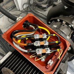 Maciej Burzyński - Otwarta, czerwona puszka elektryczna z podłączonymi kablami o różnych kolorach, widoczne złączki i narzędzia. Element instalacji elektrycznej pompy przemysłowej.