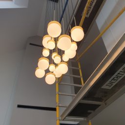 Maciej Burzyński - Nowoczesny żyrandol kulisty z żółtymi akcentami, zawieszony w wysokim pomieszczeniu, widoczny z dołu, z elementami rusztowania malarskiego.