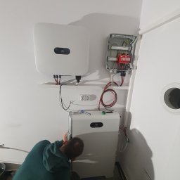 Maciej Burzyński - Instalacja paneli fotowoltaicznych w Białymstoku: monter w trakcie podłączania inwertera i akumulatora do rozdzielnicy. Widoczne kable i urządzenia.