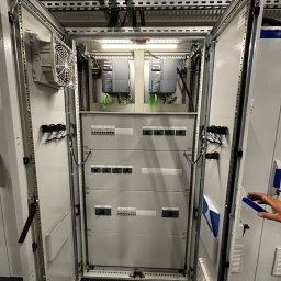 Maciej Burzyński - Otwarta szafa elektryczna z widocznymi elementami instalacji, w tym modułami sterującymi i zabezpieczeniami. Widoczne kable i przewody. Górny system podtrzymujący kable.