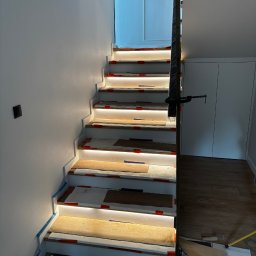 Maciej Burzyński - Montaż oświetlenia schodowego: białe schody z podświetleniem LED, widoczne elementy montażowe, zabezpieczenia, fragment podłogi i balustrady. Prace w toku.