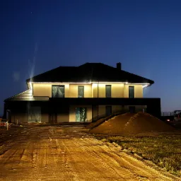 Nowoczesny, dwukondygnacyjny dom w nocnej scenerii z oświetleniem zewnętrznym, widoczne prace ziemne i sprzęt budowlany w tle, niebo ciemnoniebieskie.