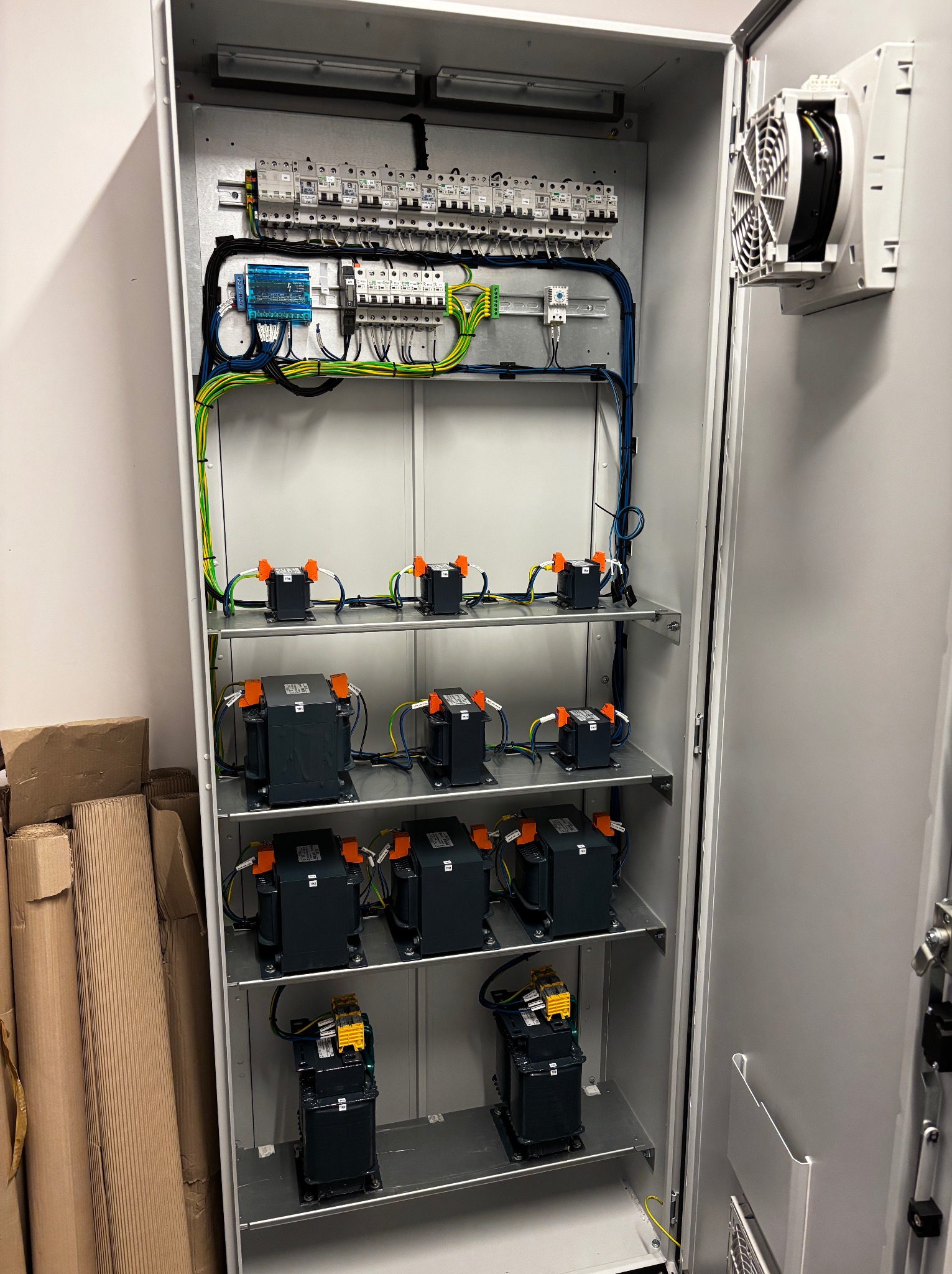 Otwarta szafa elektryczna z licznymi transformatorami, bezpiecznikami i okablowaniem. Widoczny wentylator chłodzący i elementy sterujące.