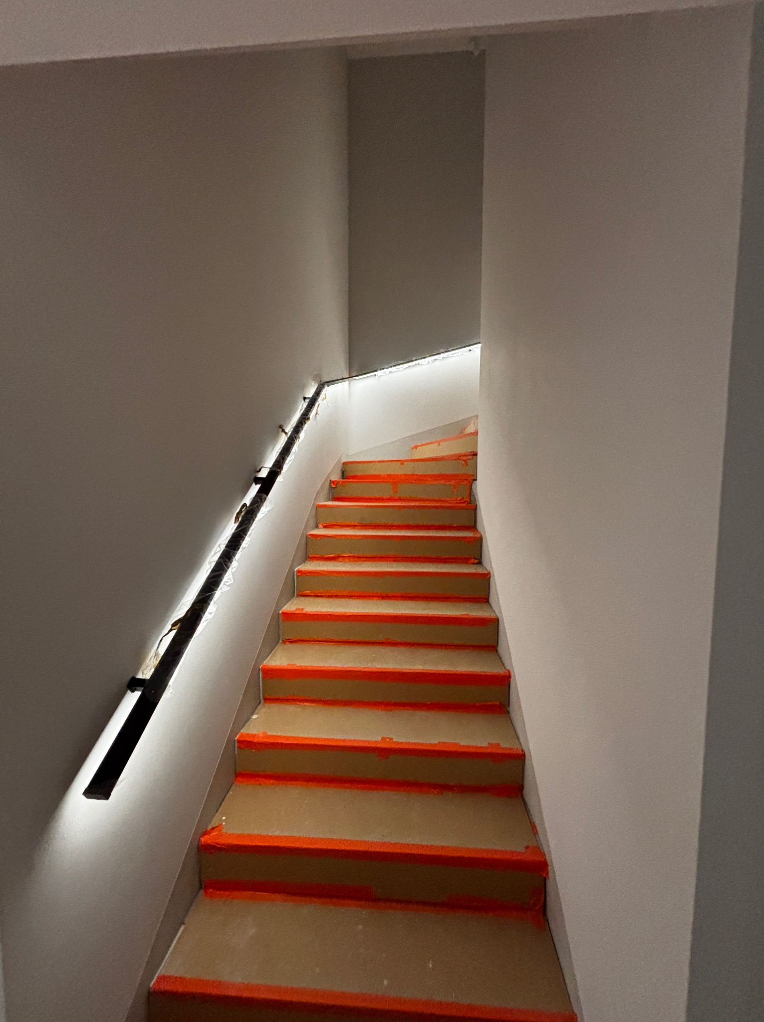 Schody z zabezpieczonymi stopniami i podświetloną poręczą, oświetlenie LED na ścianie. Widok z dołu, w trakcie prac wykończeniowych.