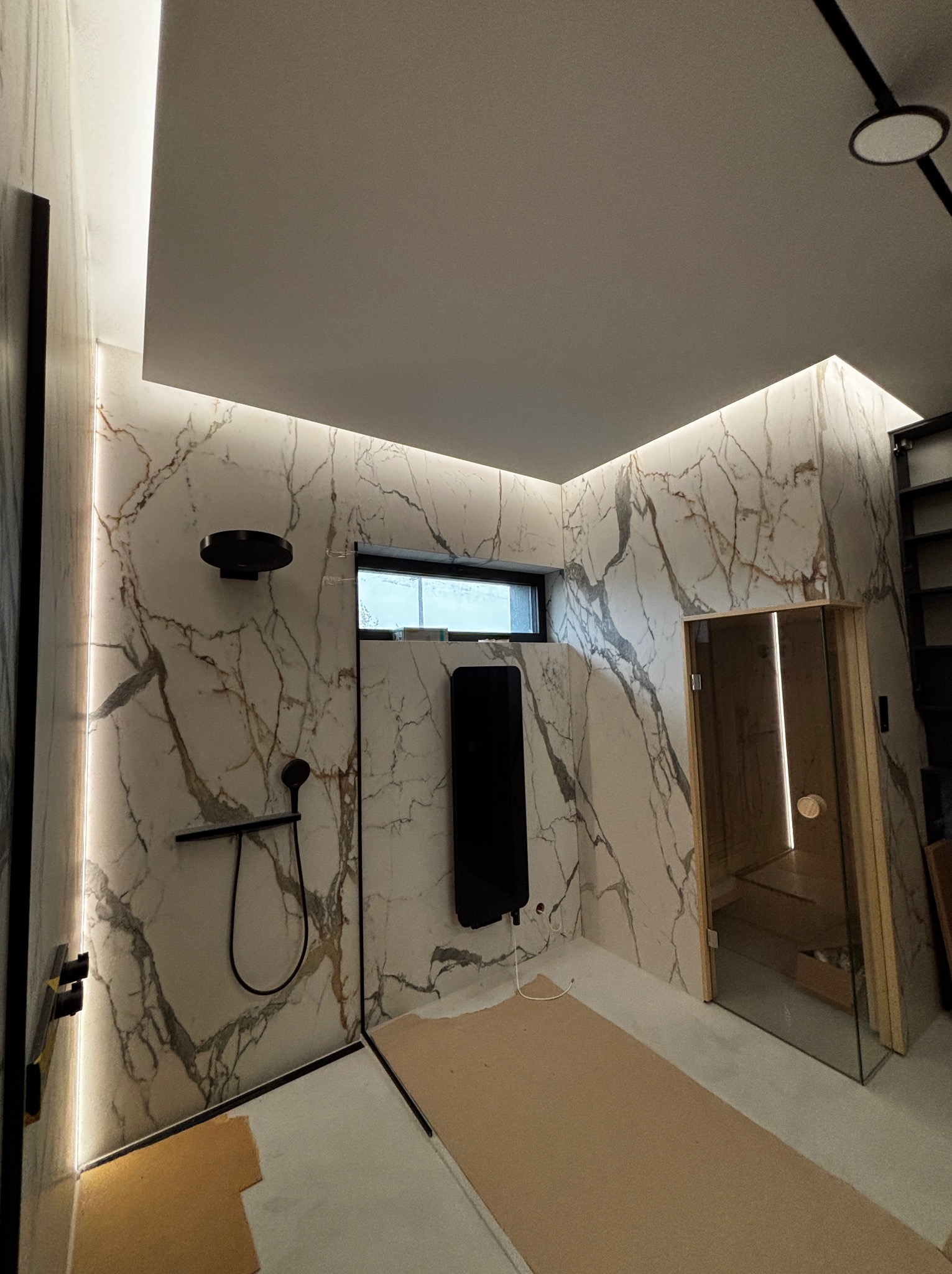Nowoczesna łazienka w trakcie wykańczania: marmurowe ściany z oświetleniem LED, czarna armatura prysznicowa, grzejnik i przeszklona sauna. Podłoga zabezpieczona kartonem.