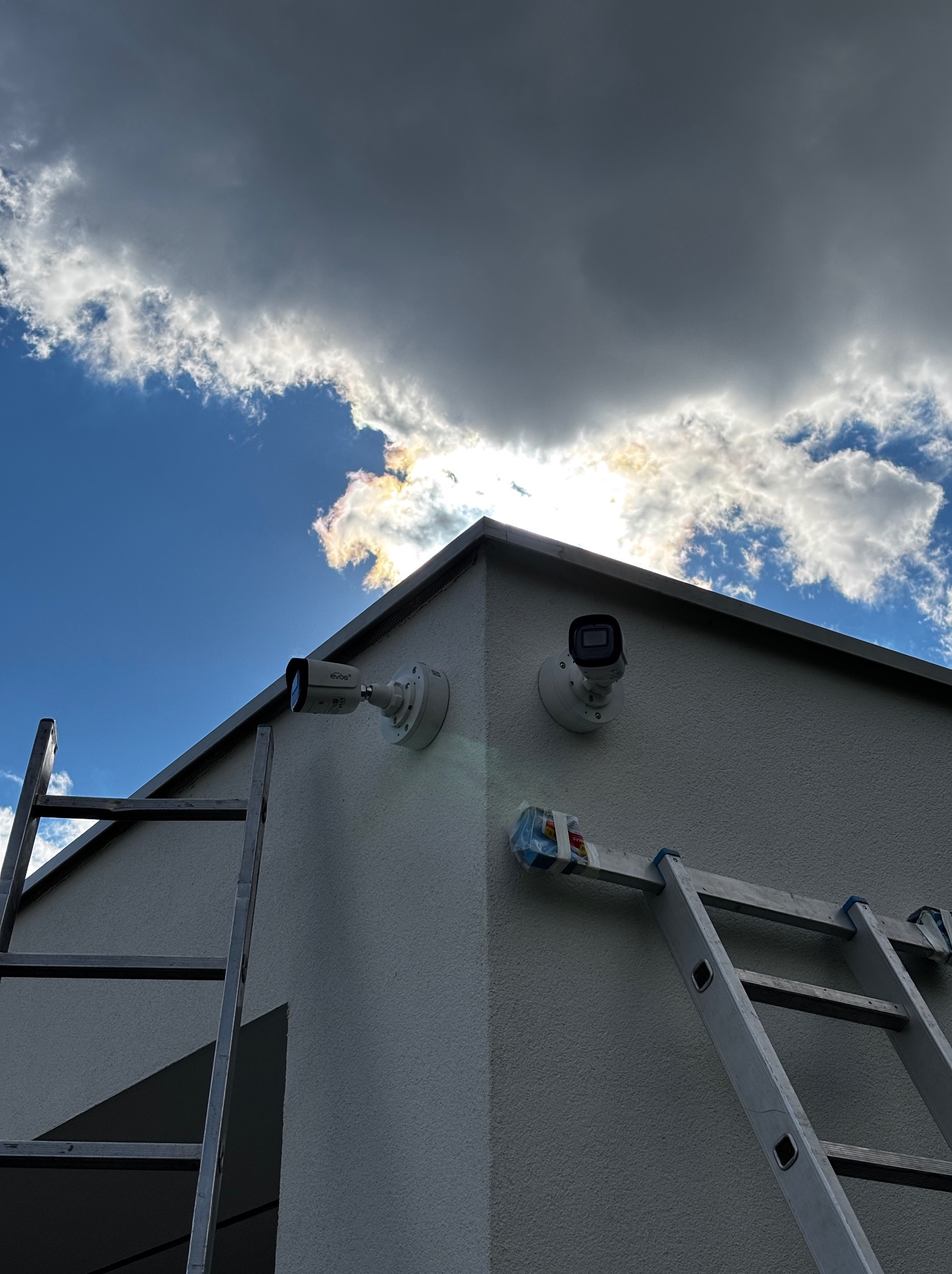 Dwie kamery monitoringu zamontowane na elewacji budynku, widoczne drabiny, niebo z chmurami w tle. Kątowe ujęcie, światło słoneczne prześwitujące przez chmury.