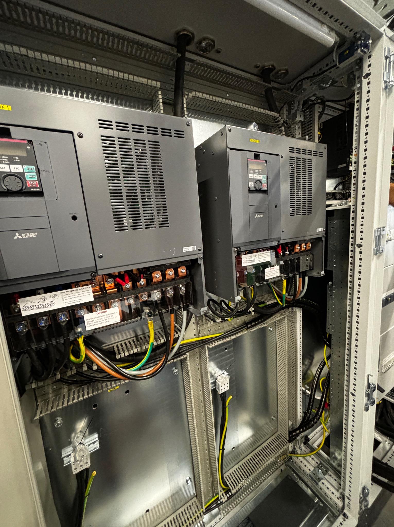 Dwa falowniki Mitsubishi Electric w szafie sterowniczej, widoczne okablowanie, złącza i etykiety. Układ wewnątrz szafy, szare obudowy falowników z wentylacją.
