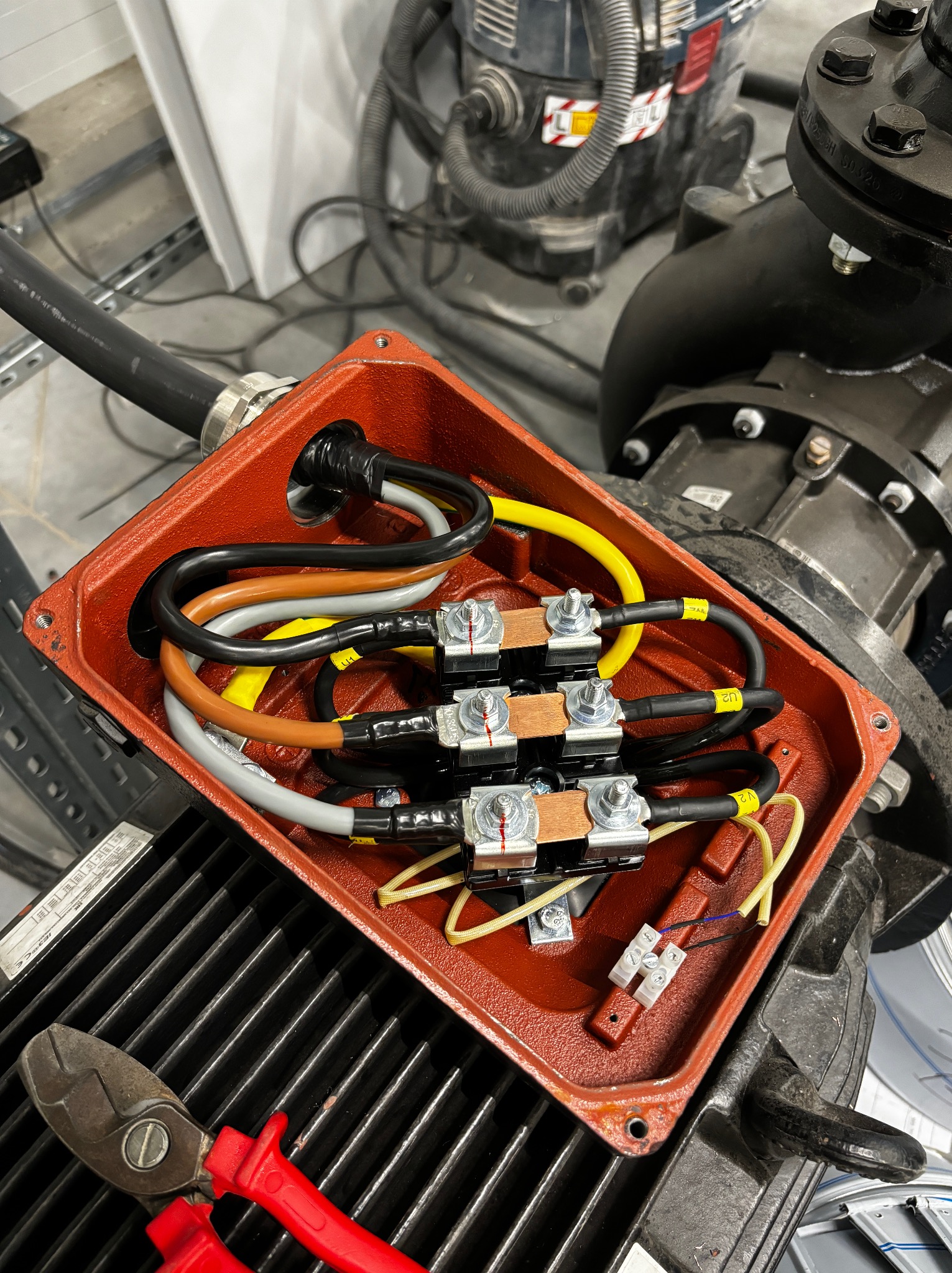 Otwarta, czerwona puszka elektryczna z podłączonymi kablami o różnych kolorach, widoczne złączki i narzędzia. Element instalacji elektrycznej pompy przemysłowej.