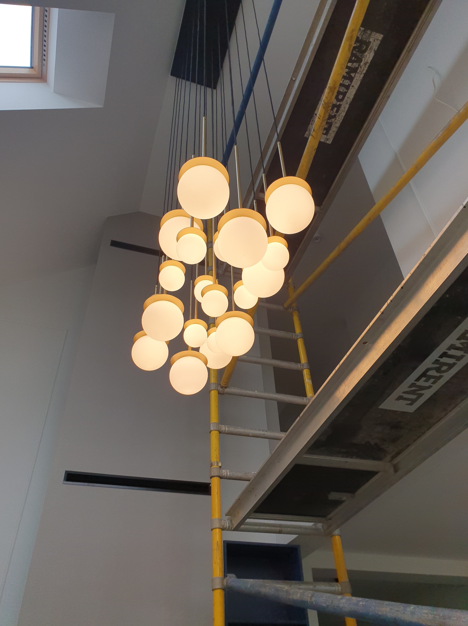 Nowoczesny żyrandol kulisty z żółtymi akcentami, zawieszony w wysokim pomieszczeniu, widoczny z dołu, z elementami rusztowania malarskiego.