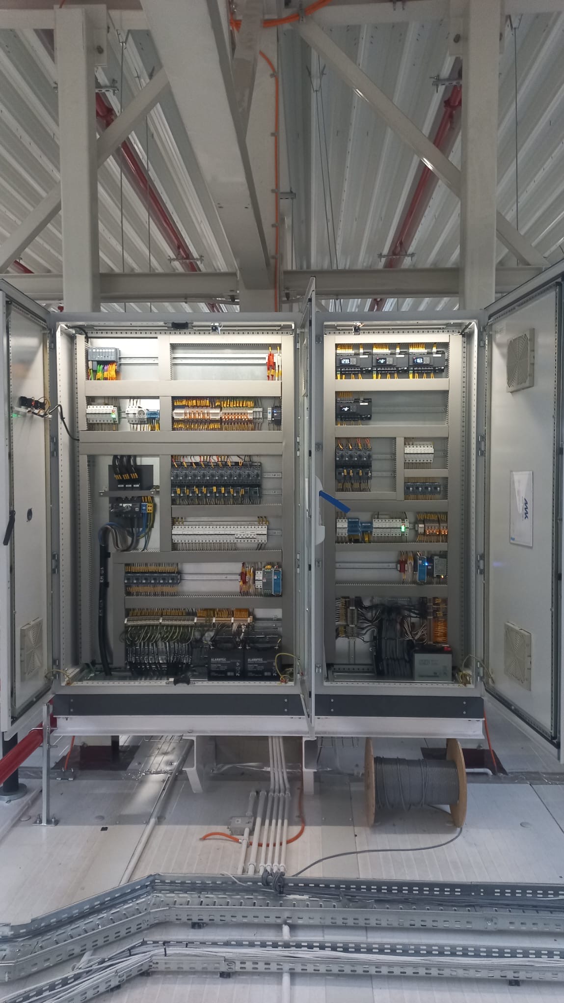 Otwarta szafa sterownicza z widocznym okablowaniem, elementami elektrycznymi i zabezpieczeniami. Na podłodze kable w korytkach i szpula kabla. Widok z przodu.