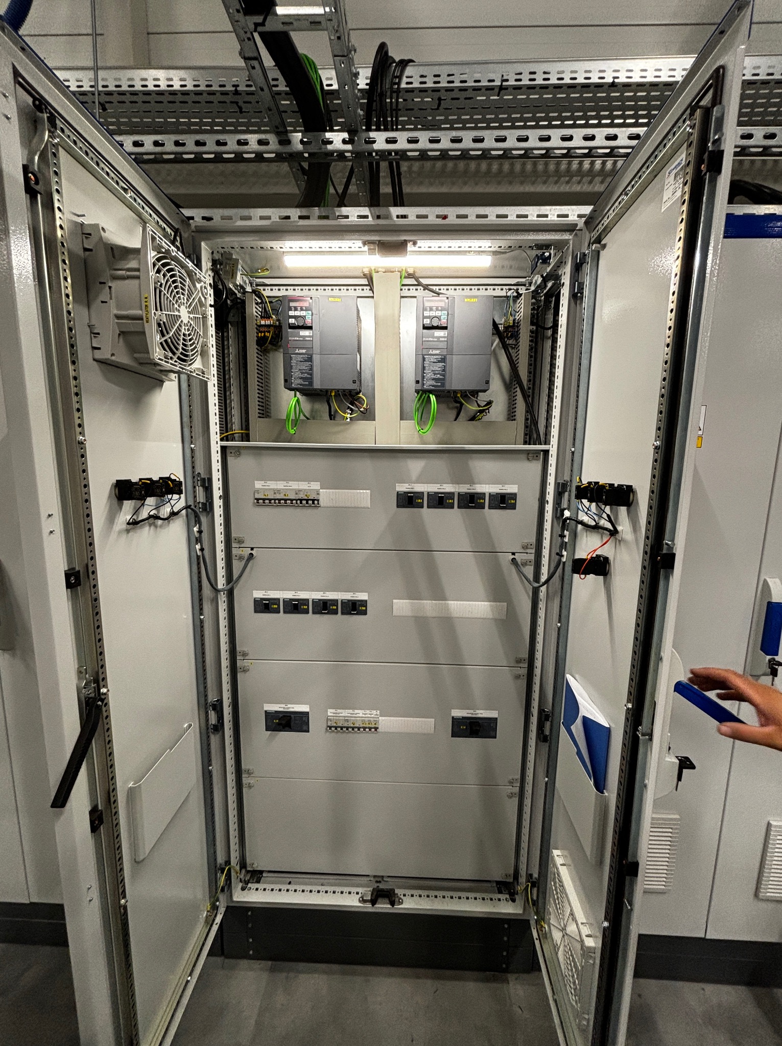 Otwarta szafa elektryczna z widocznymi elementami instalacji, w tym modułami sterującymi i zabezpieczeniami. Widoczne kable i przewody. Górny system podtrzymujący kable.