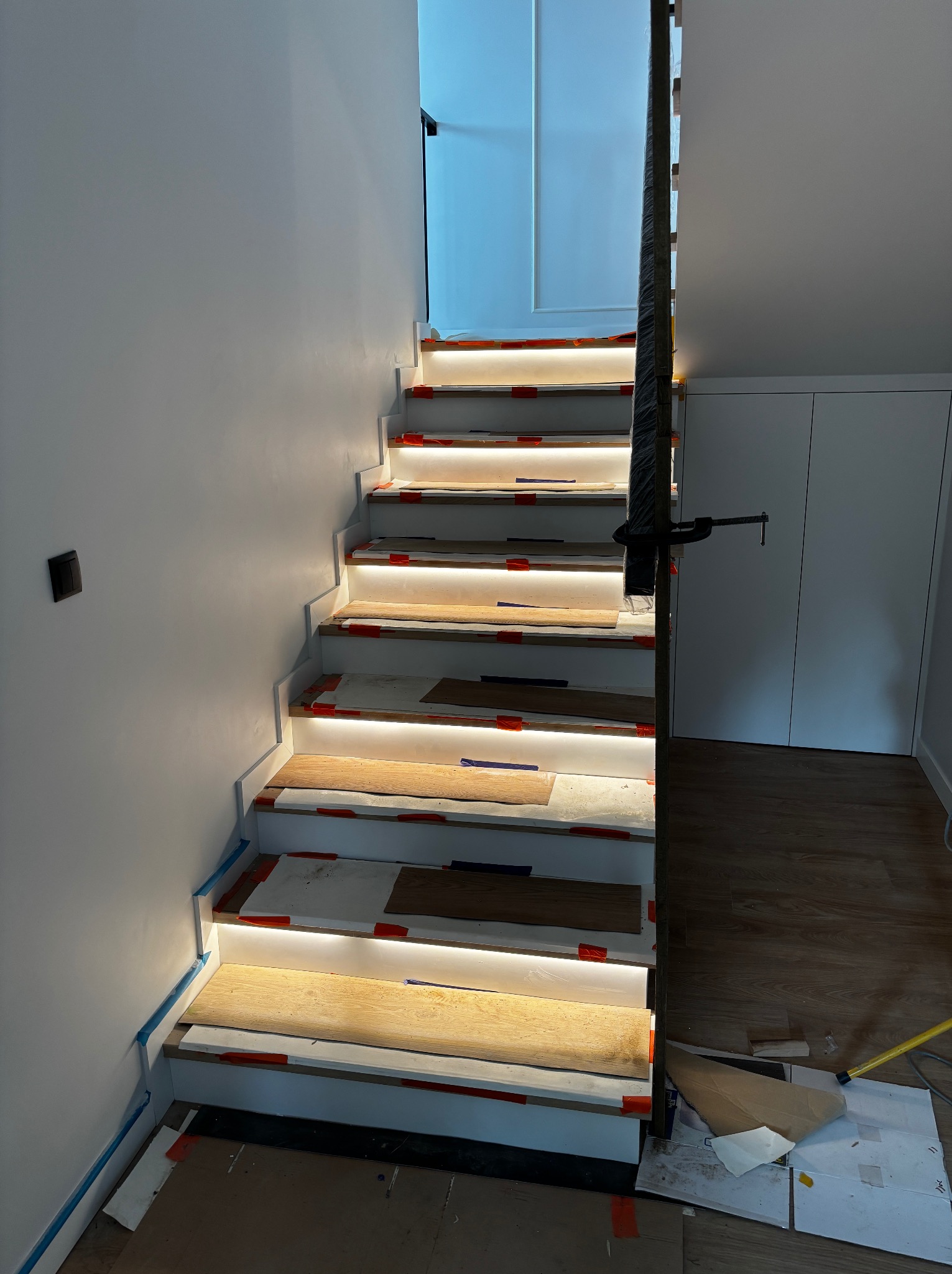 Montaż oświetlenia schodowego: białe schody z podświetleniem LED, widoczne elementy montażowe, zabezpieczenia, fragment podłogi i balustrady. Prace w toku.