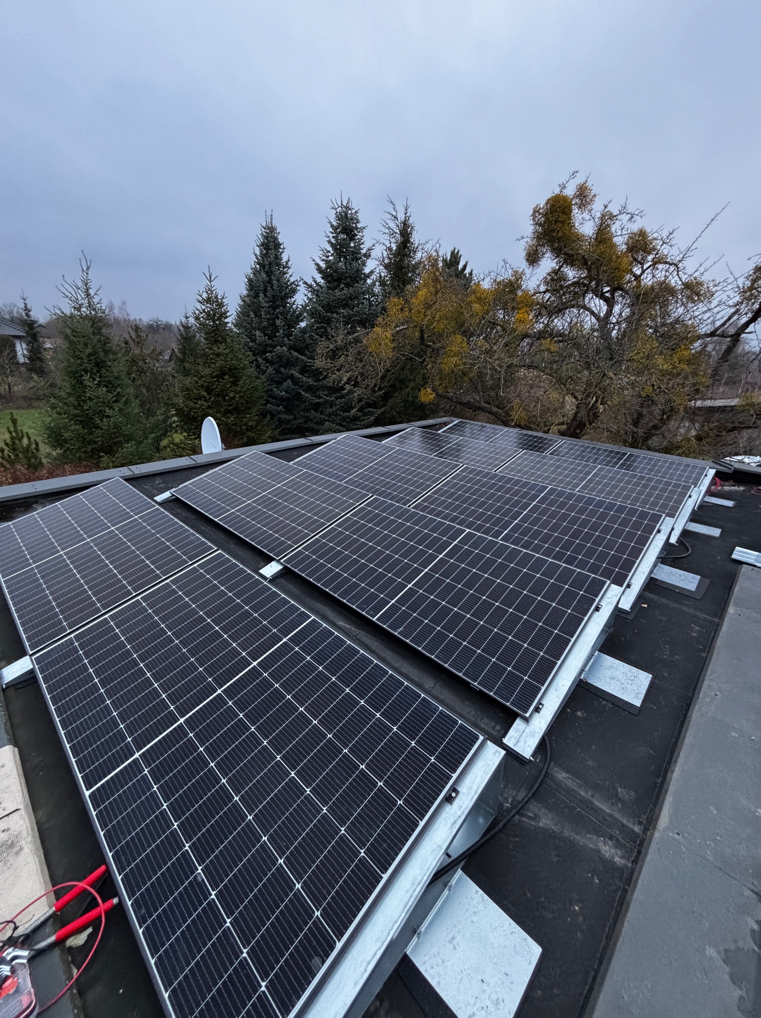 Panele słoneczne na dachu z widocznymi przewodami i elementami montażowymi, tle drzewa iglaste i budynek w pochmurny dzień. Widok z góry, ukończona instalacja.