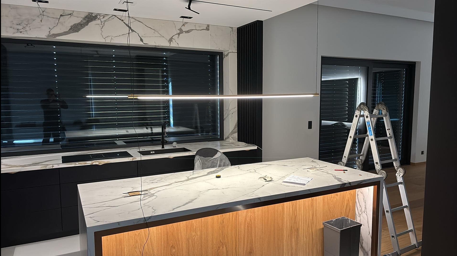 Nowoczesna kuchnia z marmurowym blatem i minimalistycznym oświetleniem LED. Widoczna drabina sugeruje prace wykończeniowe lub instalacyjne.