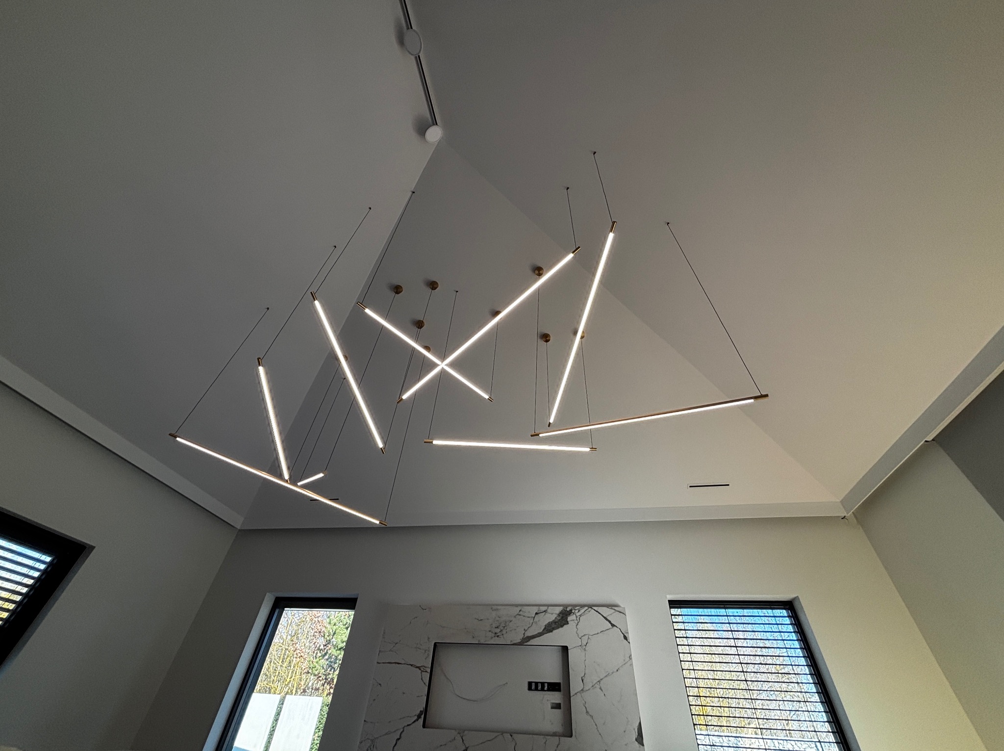 Nowoczesne oświetlenie sufitowe z podłużnych lamp LED na cienkich linkach w jasnym wnętrzu. Widok z dołu, podkreślający geometryczny design i minimalistyczny charakter.