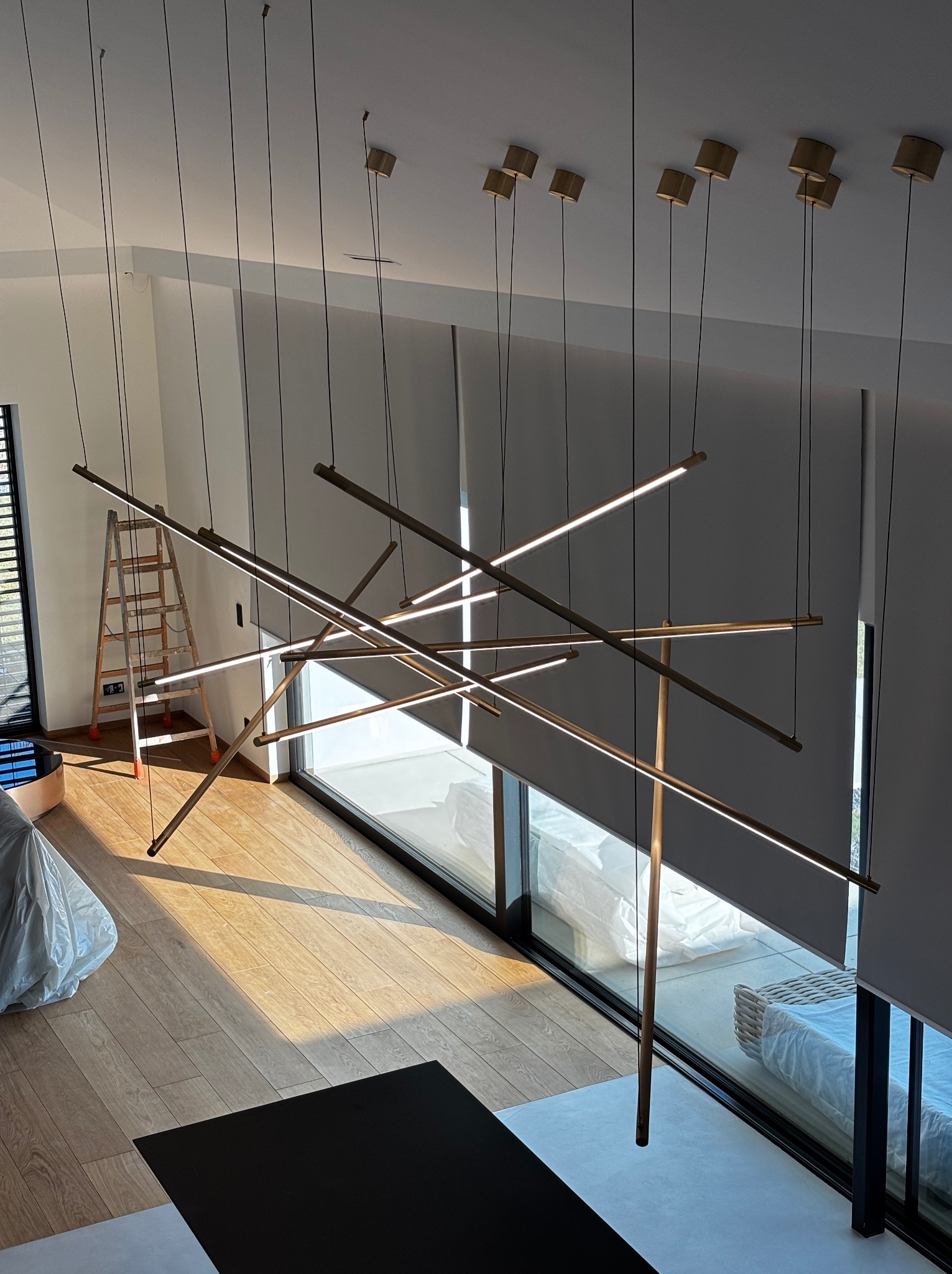 Nowoczesna lampa wisząca z elementami LED w przestronnym wnętrzu. Widoczna drabina i okna z roletami, sugerujące prace wykończeniowe. Ujęcie z góry.