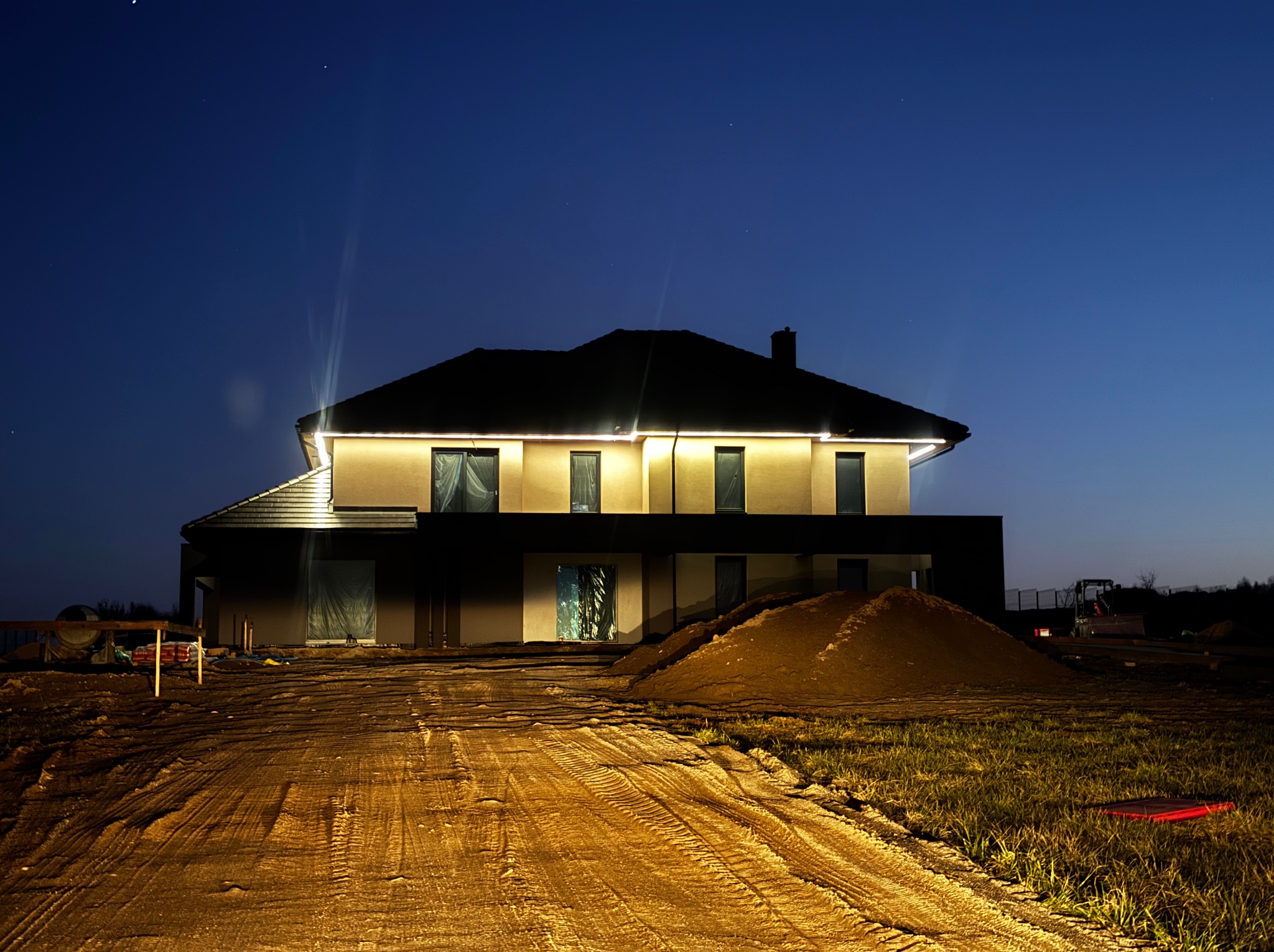 Nowoczesny, dwukondygnacyjny dom w nocnej scenerii z oświetleniem zewnętrznym, widoczne prace ziemne i sprzęt budowlany w tle, niebo ciemnoniebieskie.