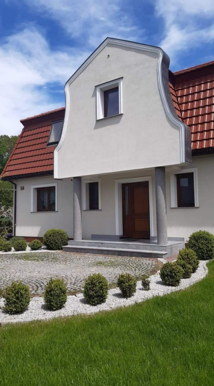 Elewacja domu jednorodzinnego w Chwaliszowie z oknami, brązowymi drzwiami wejściowymi, kolumnami i dekoracyjnym wykończeniem szczytu, otoczona trawnikiem i ozdobnymi krzewami.