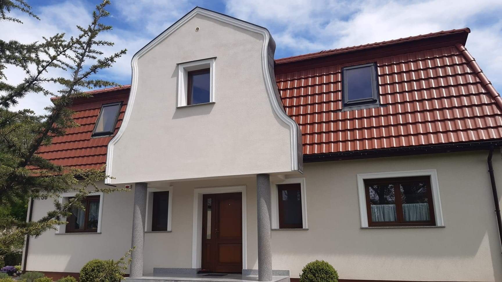 Ocieplony dom jednorodzinny z kremową elewacją, brązową dachówką i oknami, widok na front z kolumnami przy wejściu, błękitne niebo w tle.