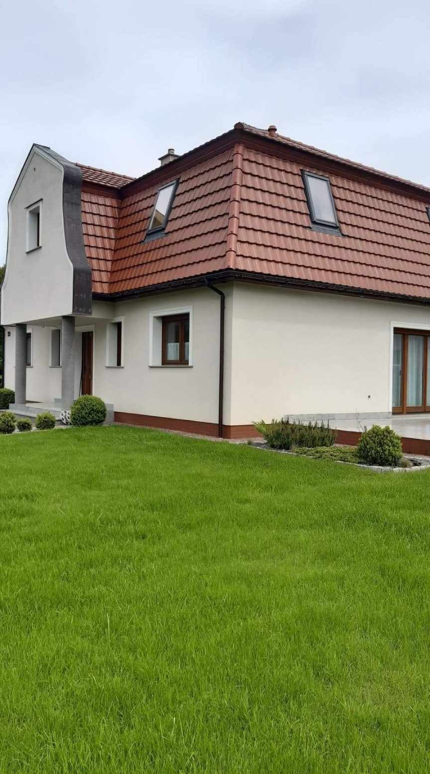 Dom jednorodzinny z beżową elewacją, czerwoną dachówką i brązowymi oknami, widok z przodu na zadbany trawnik.