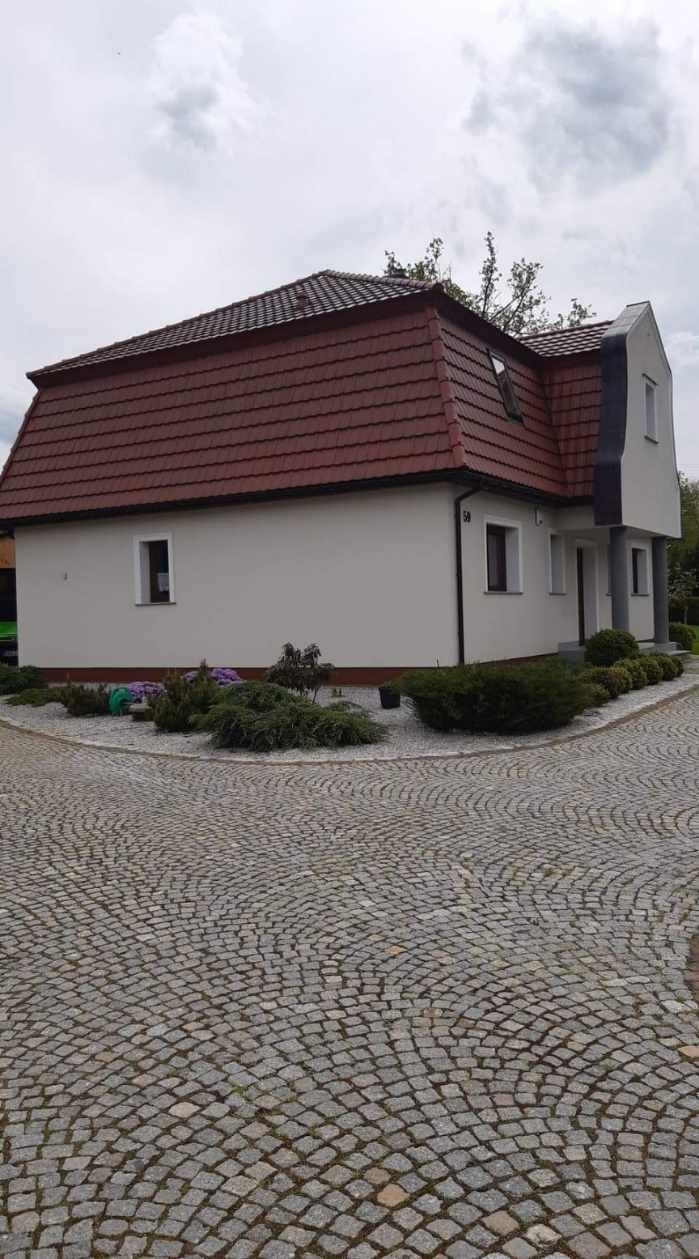 Biały dom z czerwoną dachówką, widok z zewnątrz na elewację i otoczenie z kostki brukowej i niskich krzewów.